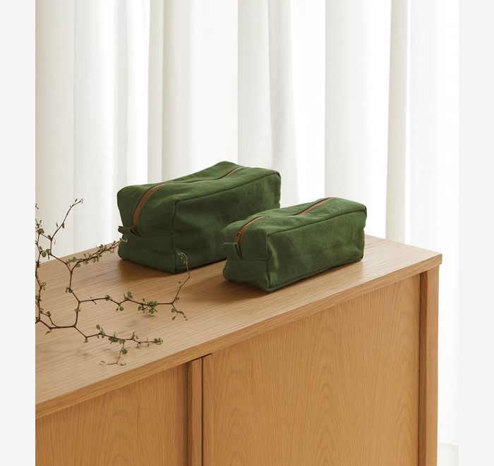 Two green toiletry bags on a wooden surface with white curtains in the background