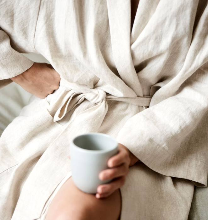 Person wrapped in a beige robe holding a white mug, sitting on a bed.