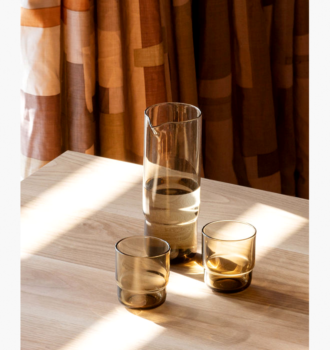 Clear glass pitcher and two glasses on a wooden surface with a patterned curtain in the background.