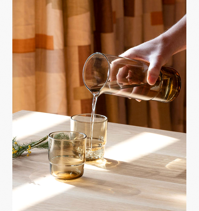 Hand pouring liquid from a glass bottle into two glasses on a wooden table with a warm-toned curtain in the background.