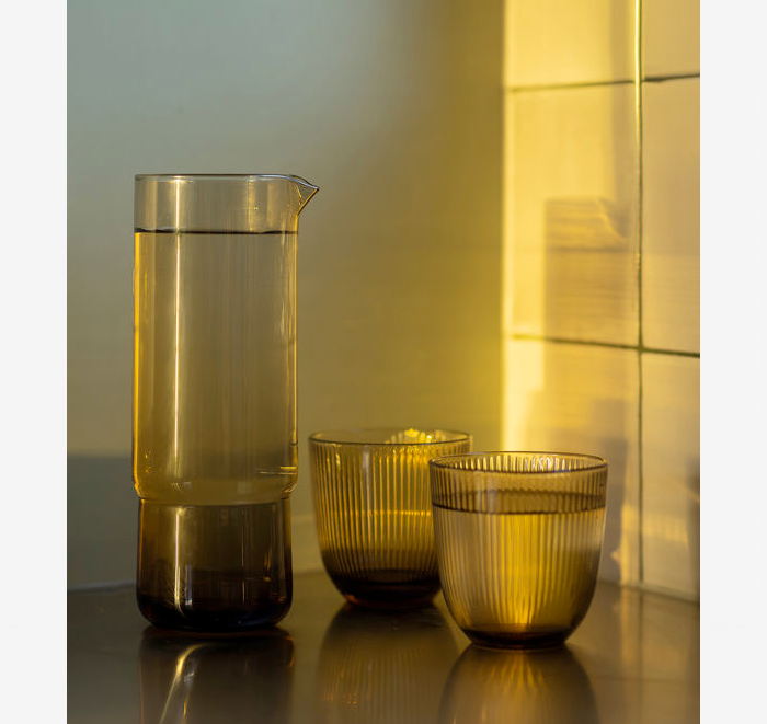 Set of glassware including a pitcher and two tumblers on a reflective surface with a tiled wall background.